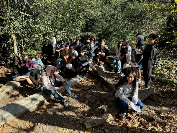 IVA High students sit on ground and logs in a forest while on outdoor field trip.