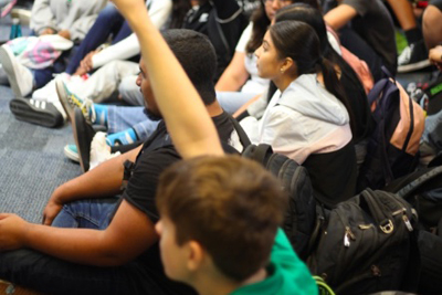 High Schoool students sitting on floor. One has a hand raised.