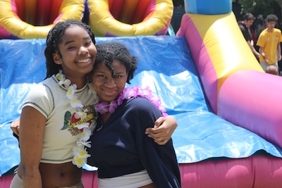 Two girls stand with arms around each other in front of a colorful bounce house slide