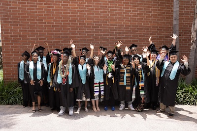 Group of IVA High seniors in cap and gown with hands in the air celebrating.