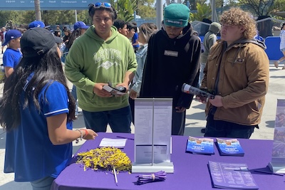 3 Students stand at table covered with purple cloth and brochures and listen to woman speaking