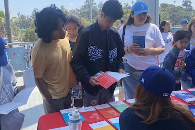 3 students look at brochures laid out on table and listen to woman behind the table speaking to them.