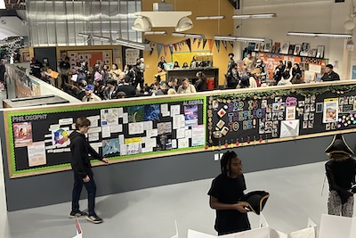  IVA High community members gather in the school to learn about student exhibitions. There are people milling around looking at colorful displays in a large open room with dividers. 