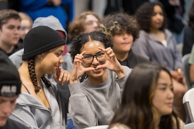 Several IVA students in chairs for assembly with one girl in glasses holding her hands up as if she’s taking a picture.