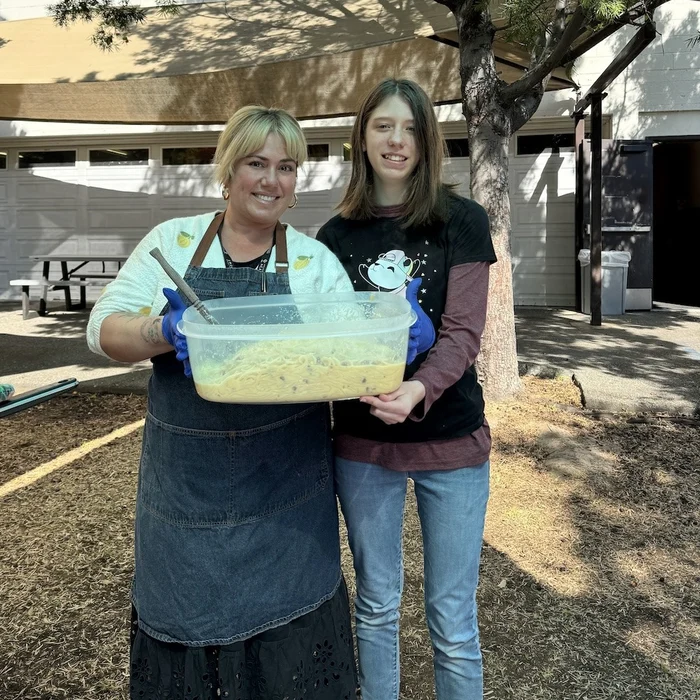 IVA High teacher, Ollie Cigliano, and a student pose together for a photo holding a large bowl of food they prepared. They are standing outside under an awning.