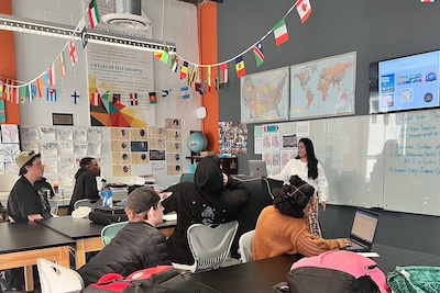 IVA High students and teacher in a classroom. The room has tall ceilings, bright colors, modern furniture and flags of the world's countries. There is a digital screen and white boards with writing.