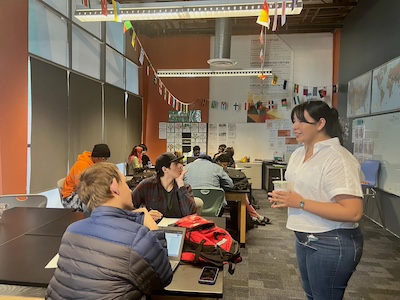 An IVA High teacher speaks with a student at a table in a classroom. Other students in the classroom are working independently. 