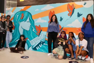 Teachers and students at IVA High stand in front of a colorful mural of a woman reading. The mural was created by students in collaboration with CreateNow. 