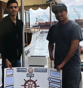 IVA High students stand with a poster presentation titled Maritime Preservation Trust. They are standing at the Long Beach Port. There is water and a ship in the background. 