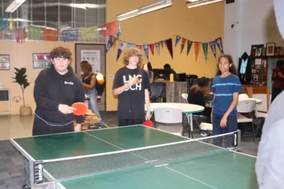 Students at IVA High play pingpong while another student looks on. They are in a large room with colorful walls and college banners decorating the walls.
