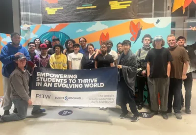 Students and staff at IVA High gather for a photo while standing in front of a brightly colored mural in the school building. They are holding a banner that reads “Empowering students to thrive in an evolving world.”