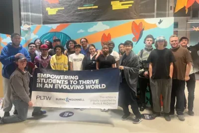 Students and staff at IVA High gather for a photo while standing in front of a brightly colored mural in the school building. They are holding a banner that reads “Empowering students to thrive in an evolving world.”