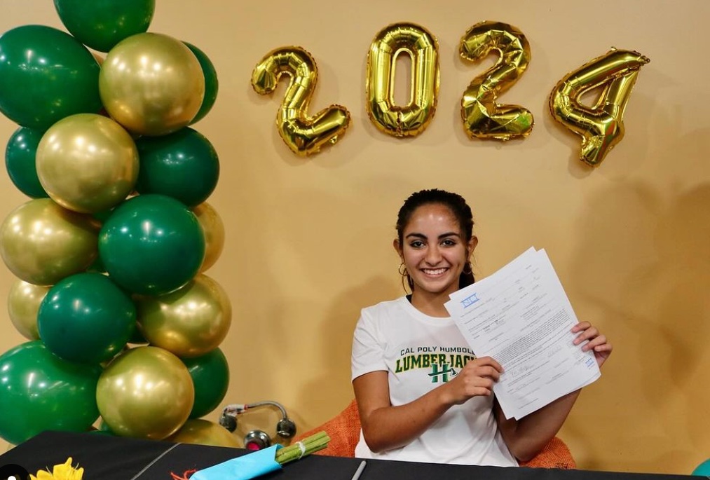 Teenager sitting in chair in front of gold and green balloons holding up a paper.
