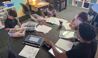 Students sit around a table with papers, pencils, and art supplies spread in front of them.