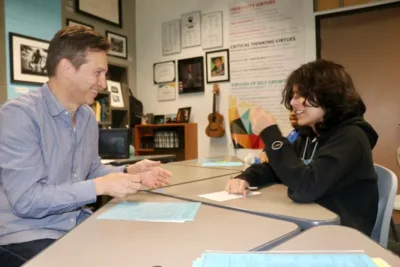 An IVA High teacher and student work together on an assignment. They are sitting across from each other at a table and both are smiling.