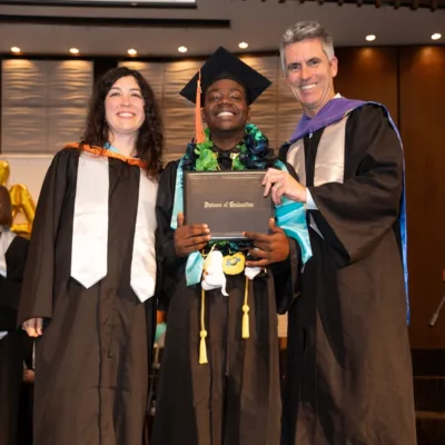 An IVA High graduate of 2023 stands between two staff members while holding his diploma. They are all wearing black robes and colorful stoles and hoods.