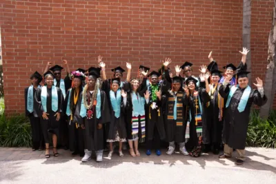IVA High graduates in caps and gowns stand against a red brick wall.