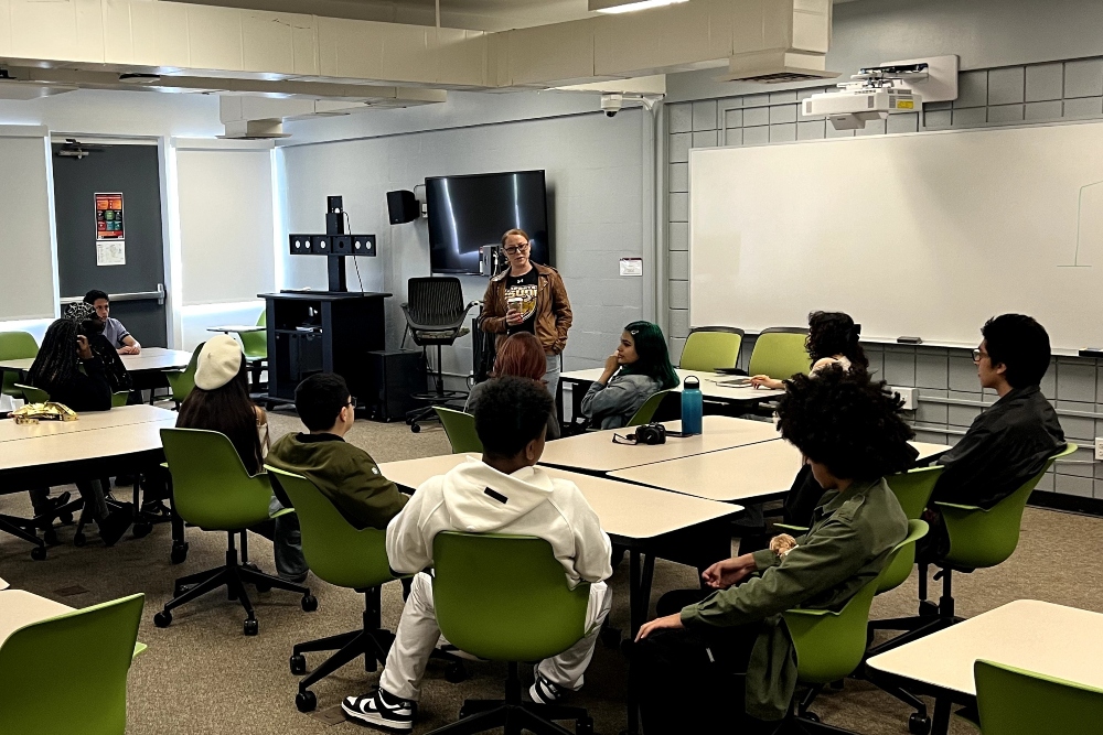 IVA High students sit in desks in a classroom while listening to a teacher.