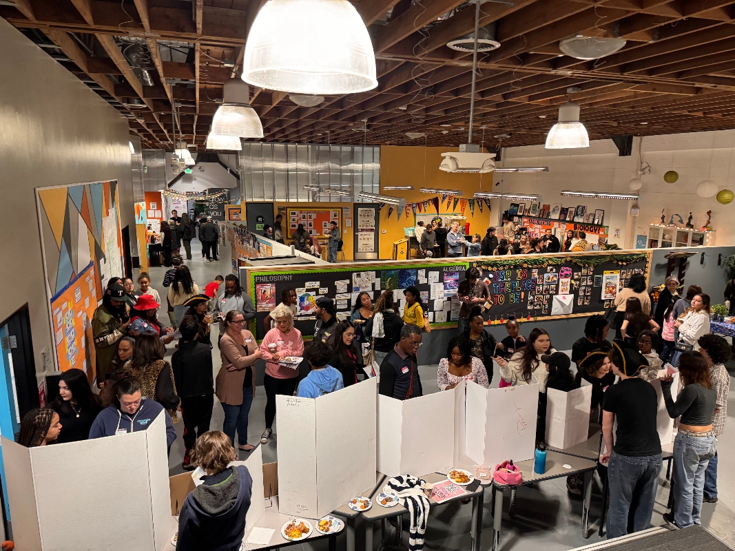 An aerial view of IVA High during the Exhibition of Learning in Long Beach. Students and families are sharing projects and discussing learning in a large open room with dividers and tables.