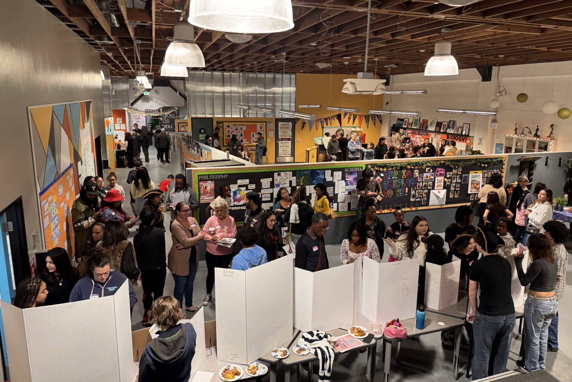 An aerial view of IVA High during the Exhibition of Learning in Long Beach. Students and families are sharing projects and discussing learning in a large open room with dividers and tables.